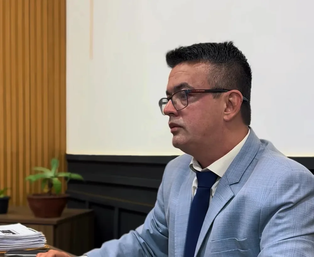 Dr. Gilbran de perfil, de terno azul-claro, trabalhando à mesa; ao fundo, a parede ripada e um vaso de planta